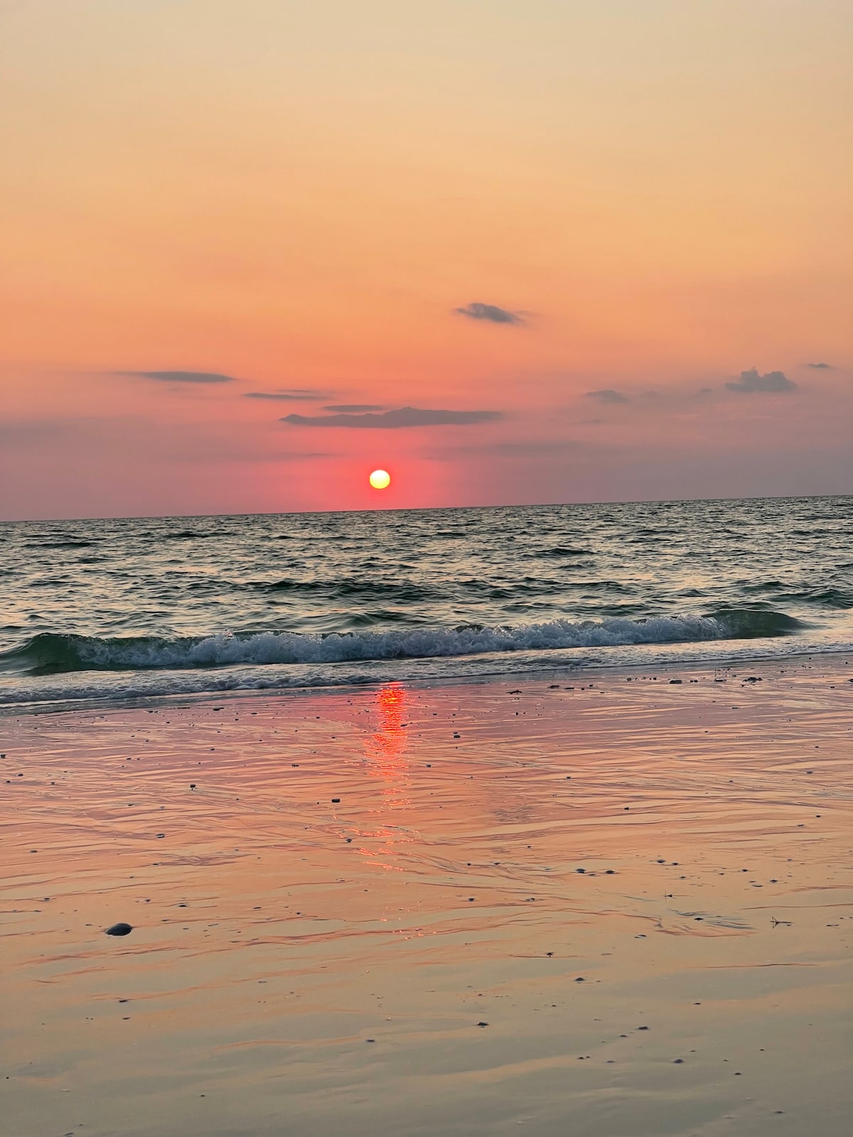 A serene view of the sun setting over the water, casting warm shades of orange and pink across the sky. Gentle waves lap at the sandy shore, reflecting the sun's glow. Soft clouds are visible in the distance, adding depth to the tranquil scene.