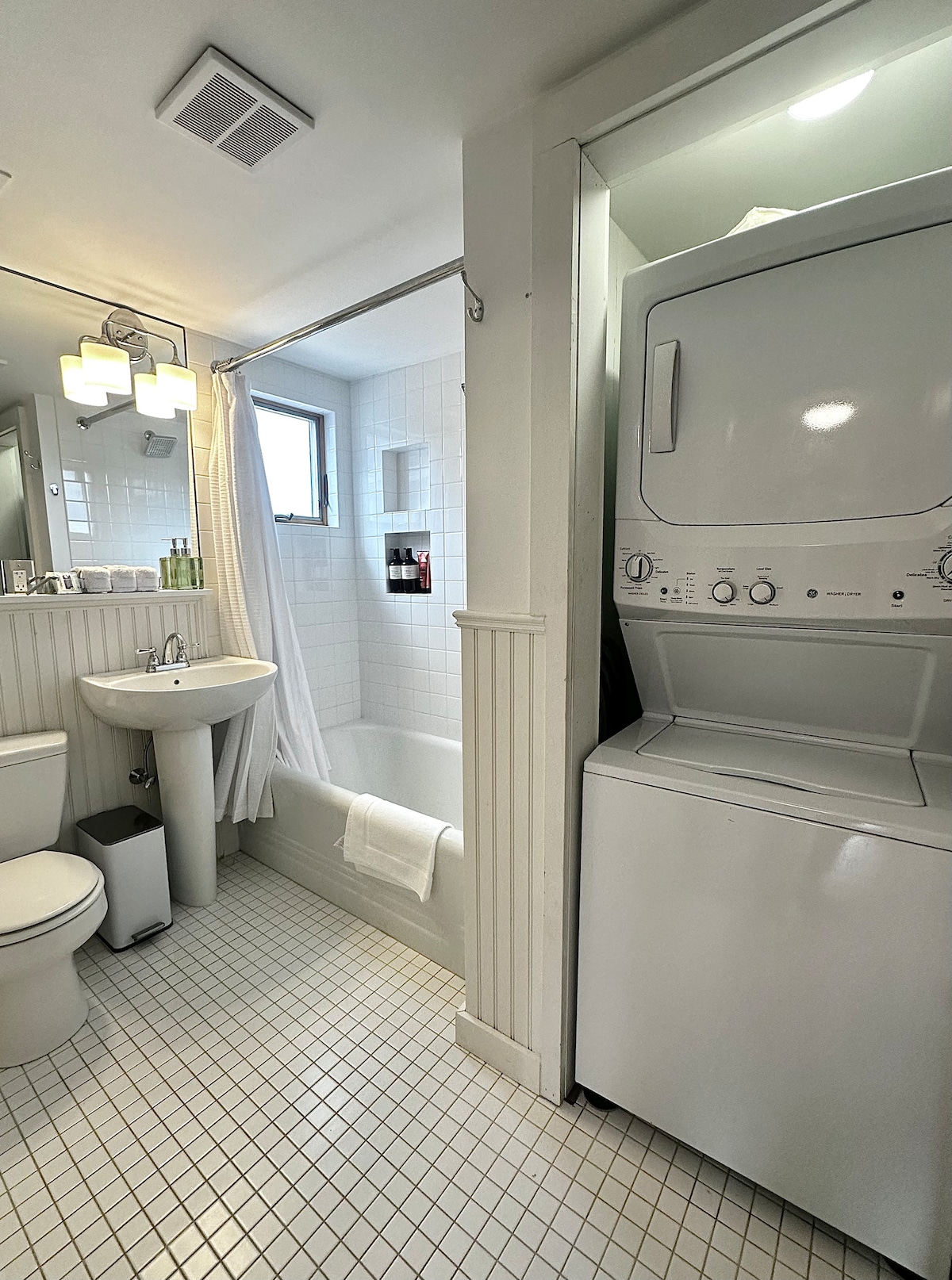 The bathroom features a clean and tidy layout with a bathtub and shower combination. A compact washer and dryer are located beside the bathtub. A sink with a mirror overhead and a toilet are also present, all set against a tiled floor.