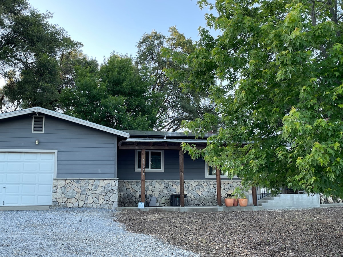 A charming exterior view of a single-story home is featured, showcasing a stone and siding facade. Lush green trees provide shade, while a gravel driveway leads to a garage. The porch is supported by wooden posts, creating a welcoming entryway.