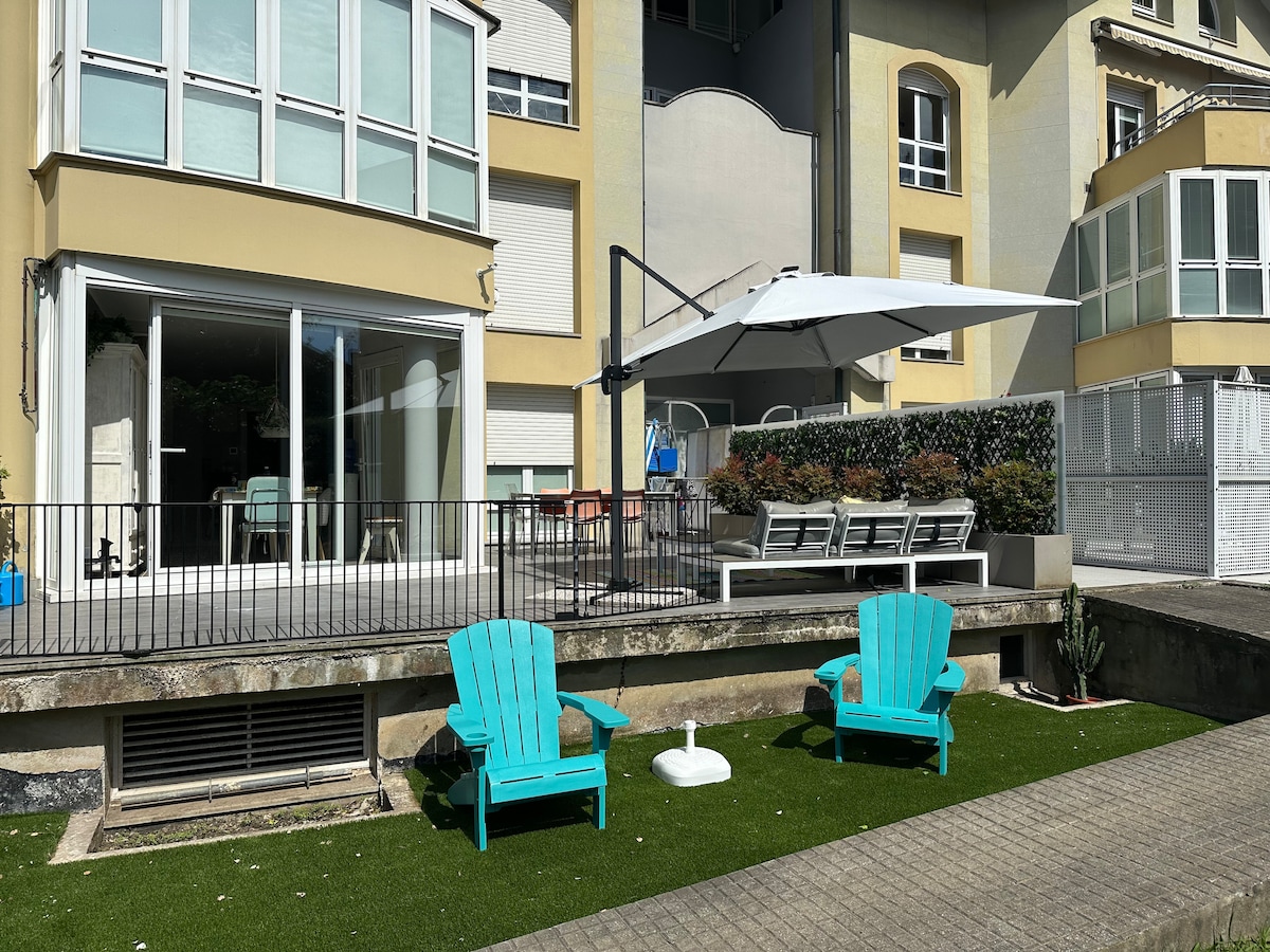 An outdoor terrace is furnished with a large shaded area and several lounge chairs, inviting relaxation. Two turquoise chairs are positioned on lush green grass, while a white umbrella offers sun protection. The terrace is framed by a modern building with expansive windows.