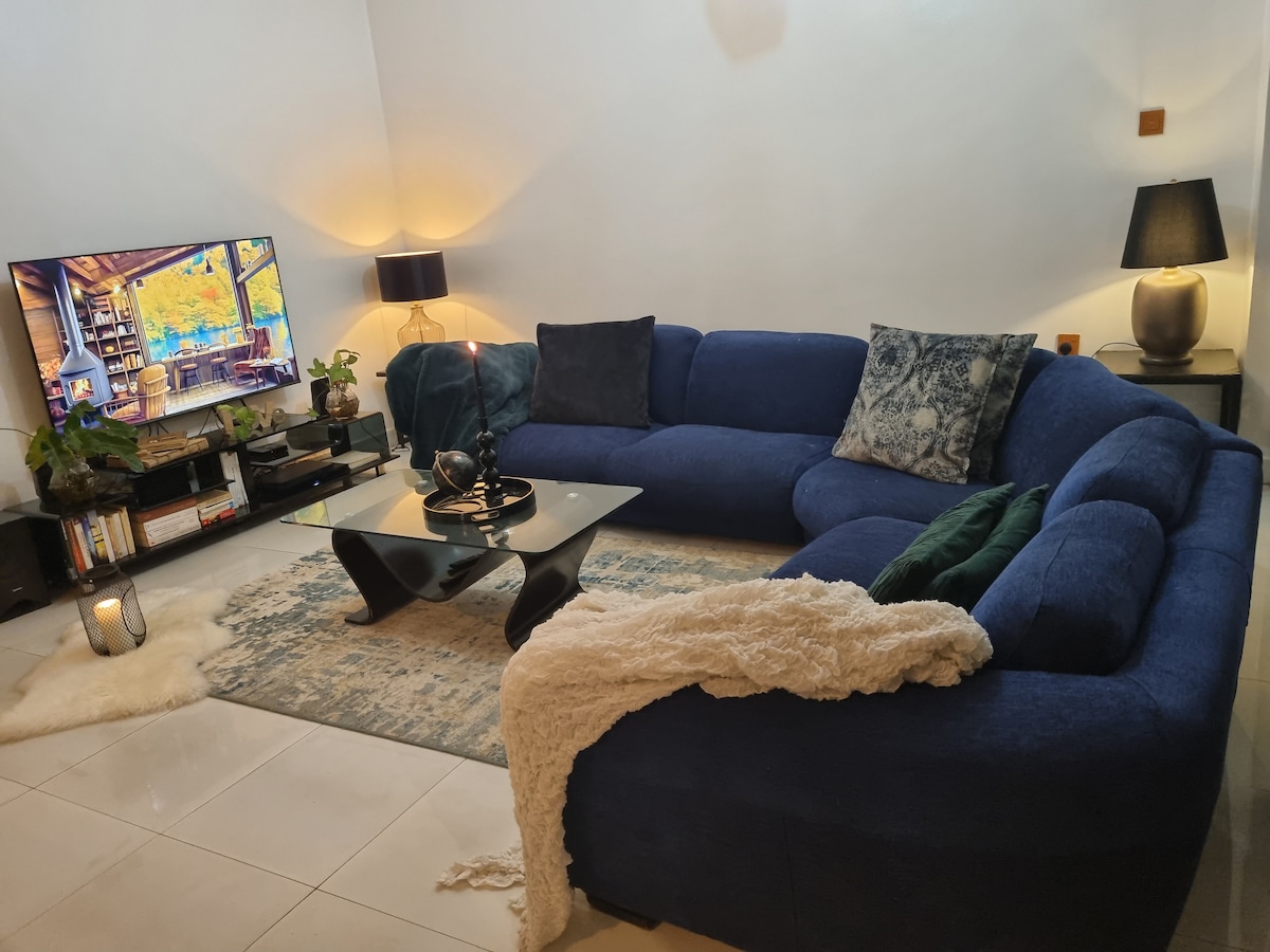 The living area is arranged with a large blue sectional sofa and a low black coffee table. A TV displays a scenic image, while a soft rug lies beneath. Ambient lighting is provided by lamps, contributing to a cozy atmosphere.