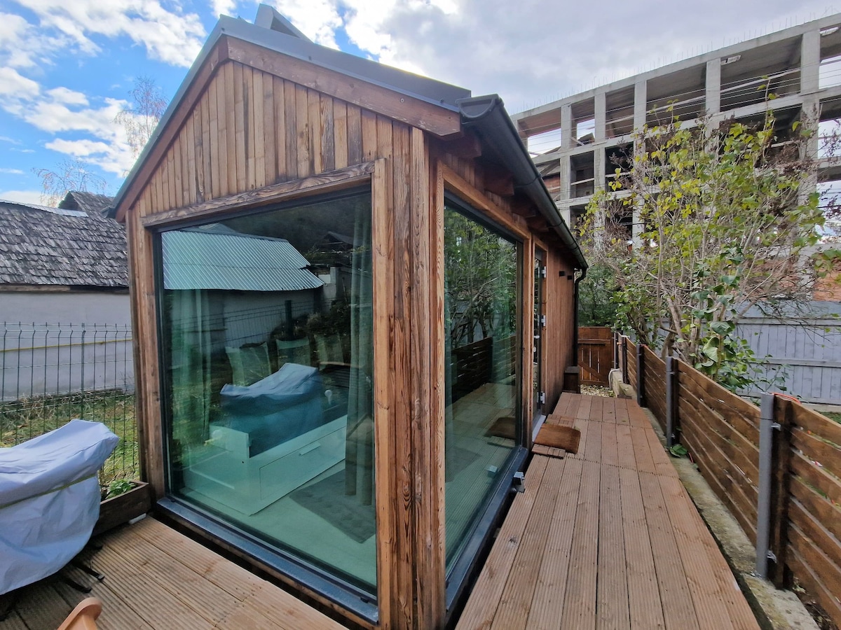 A wooden structure features large glass windows, allowing natural light to fill the space. A wooden deck extends along the exterior, providing a seamless transition between indoor and outdoor areas. The surrounding greenery contributes to a serene atmosphere.