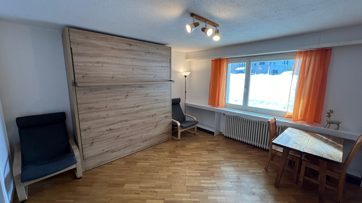 A spacious living area features a high-quality wall bed that can be folded up, providing versatility. Two comfortable chairs are positioned nearby, and a wooden dining table is set against the wall. Natural light fills the room through a large window adorned with bright orange curtains.