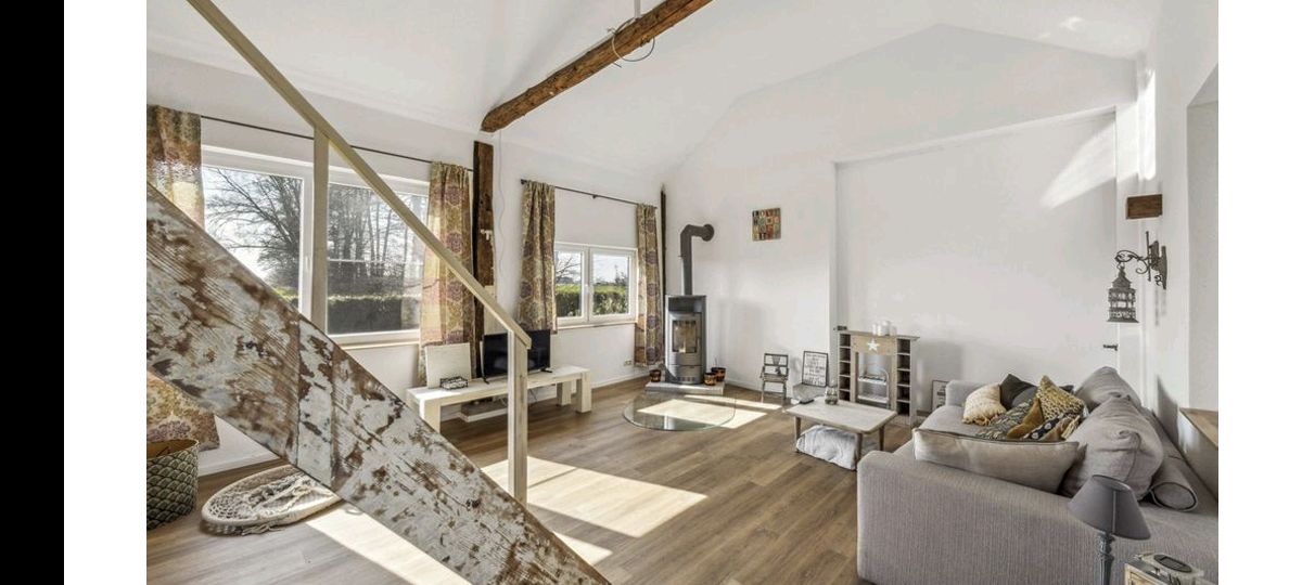 An open living area features large windows allowing natural light to fill the space. A spacious couch is positioned near a modern wood stove, accompanied by a glass coffee table. Decorative curtains frame the windows, while exposed wooden beams add character to the ceiling.