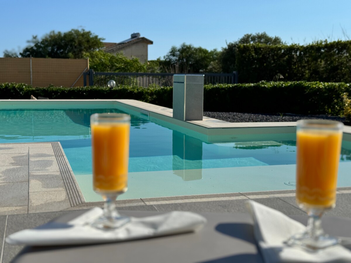 Two glasses of orange juice are placed beside a sparkling pool, reflecting the bright sunlight. The pool is surrounded by neatly trimmed hedges and a spacious deck area, creating a serene atmosphere perfect for relaxation.