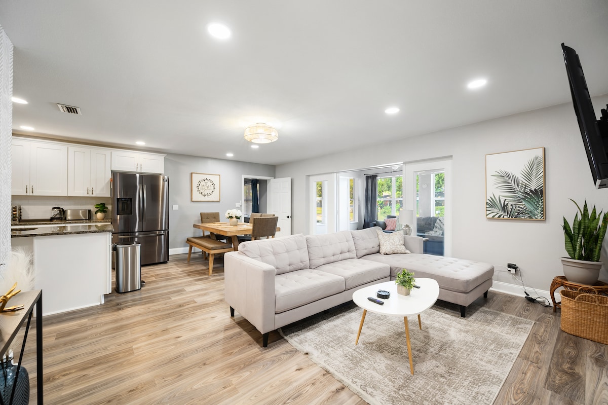 The open living area features a light-colored sectional sofa adjacent to a round coffee table. A dining table is set nearby with seating for four. Bright windows allow natural light to illuminate the space, while a large wall-mounted TV and potted plant add to the decor.