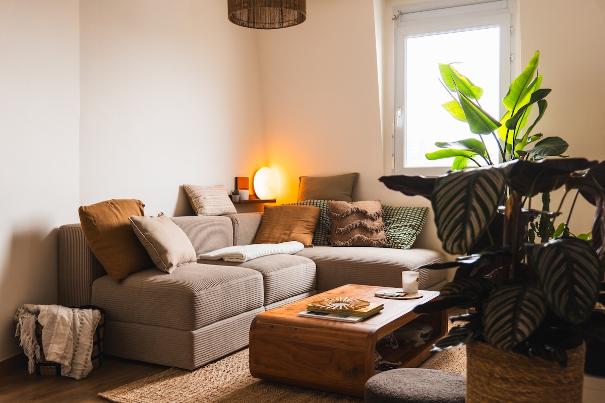 A comfortable living area is featured with a spacious sectional sofa adorned with multiple cushions in warm tones. A wooden coffee table sits in front, accompanied by a small plant. Natural light streams through a nearby window, enhancing the cozy ambiance.