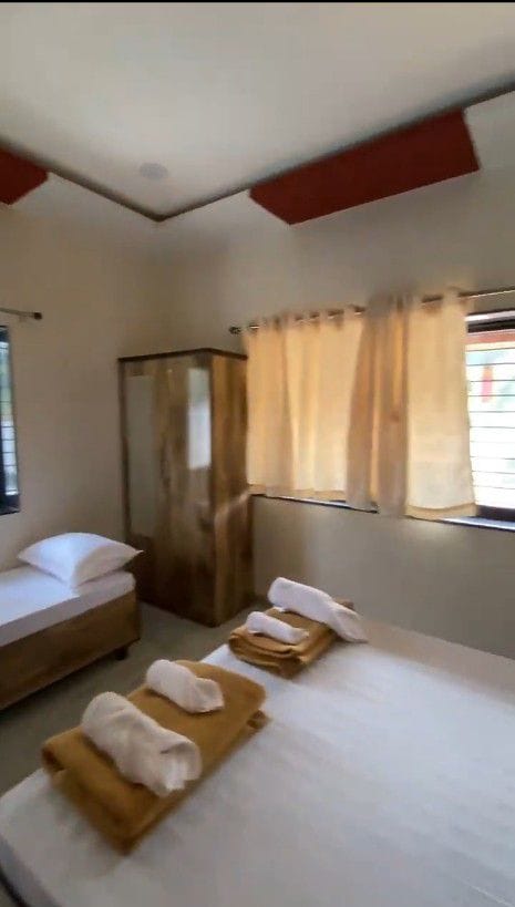 A well-lit room features a spacious bed with neatly arranged towels. A single bed is positioned alongside a wooden wardrobe and a window dressed with light, flowing curtains. The walls are adorned with a subtle, neutral hue.