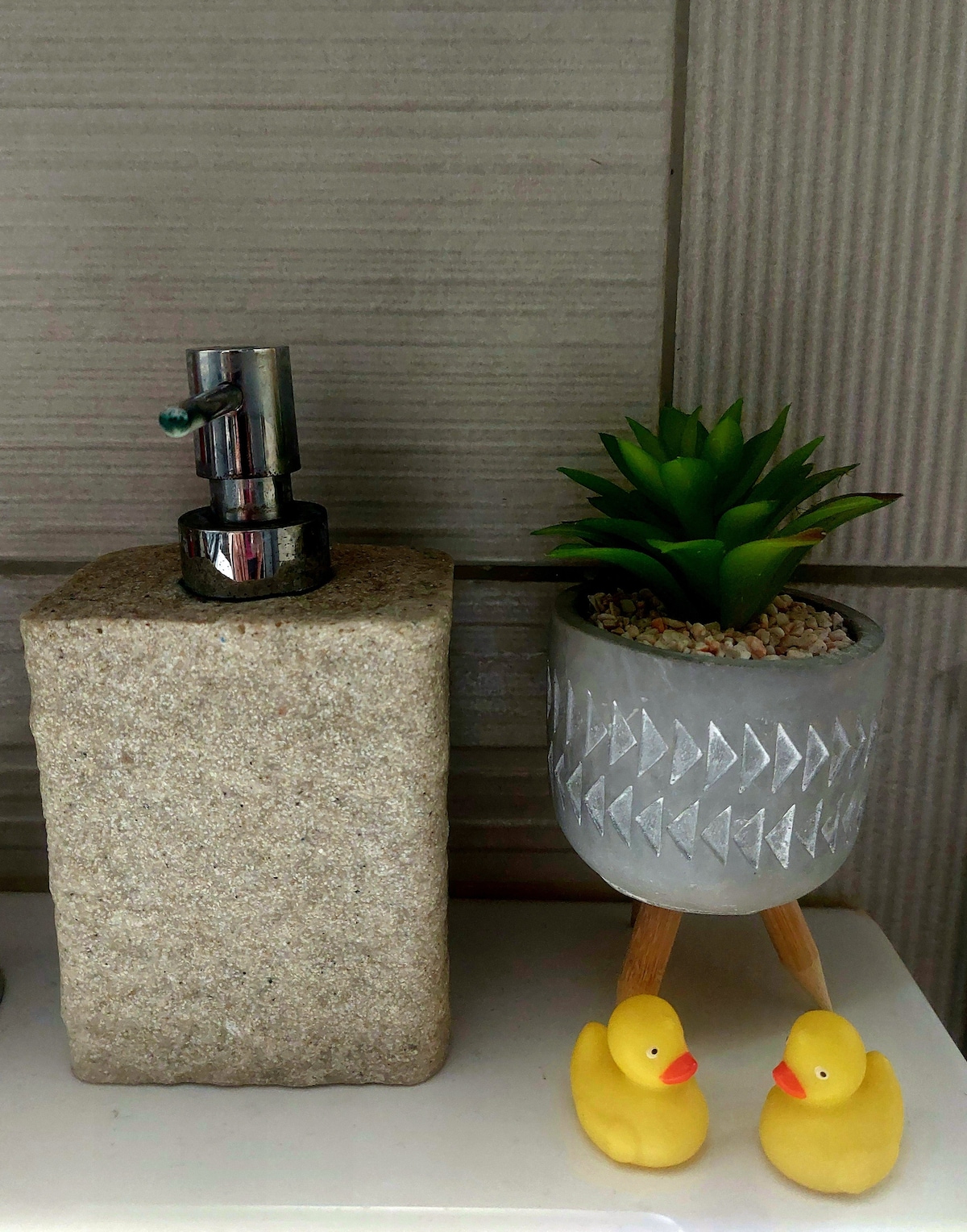 A granite soap dispenser stands next to a ceramic planter featuring a small succulent, both arranged on a bathroom counter. Below, two bright yellow rubber ducks are positioned, adding a playful touch to the decor.