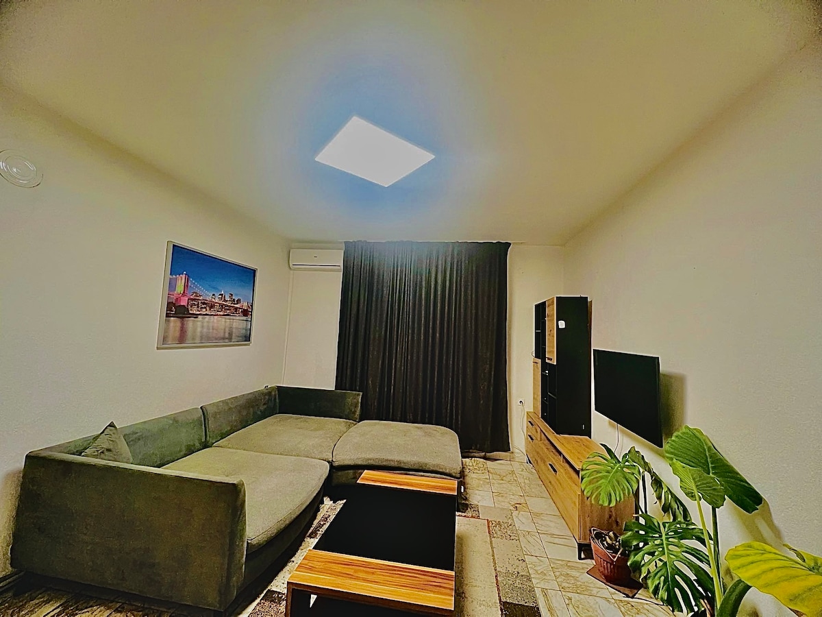 A comfortable living area features a large, L-shaped sofa and a wooden coffee table. A flat-screen television is mounted on a light-colored media console. Soft lighting is provided by a ceiling fixture, and a potted plant adds a touch of greenery. Dark curtains frame the window.
