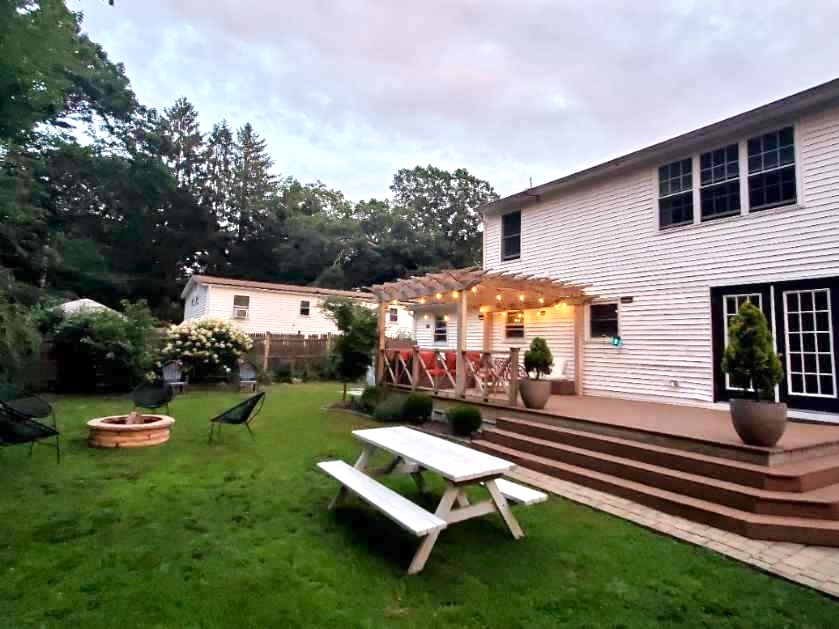 A spacious backyard features a wooden deck with outdoor seating under string lights, creating a warm ambiance. A fire pit is surrounded by seating, and a grassy area offers ample space for outdoor activities. The exterior of the house displays multiple large windows.