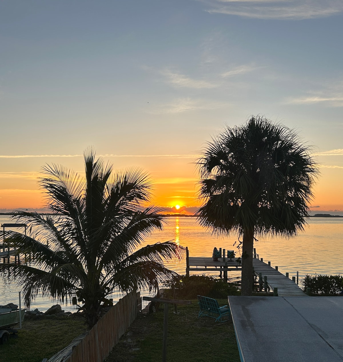 A serene view of the sunset over the Banana River is framed by vibrant palm trees. The horizon glows with warm colors, reflecting on the water's surface. A wooden dock and gentle ripples can be seen, creating a peaceful atmosphere for relaxation.