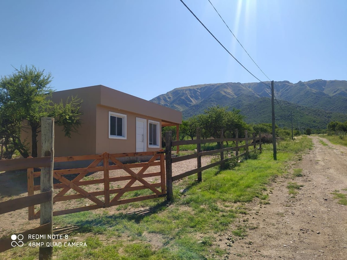 A simple mountain house is set beside a dirt path, framed by green grass and trees. The structure features a light-colored exterior and two windows, with majestic mountains rising in the background beneath a clear blue sky.