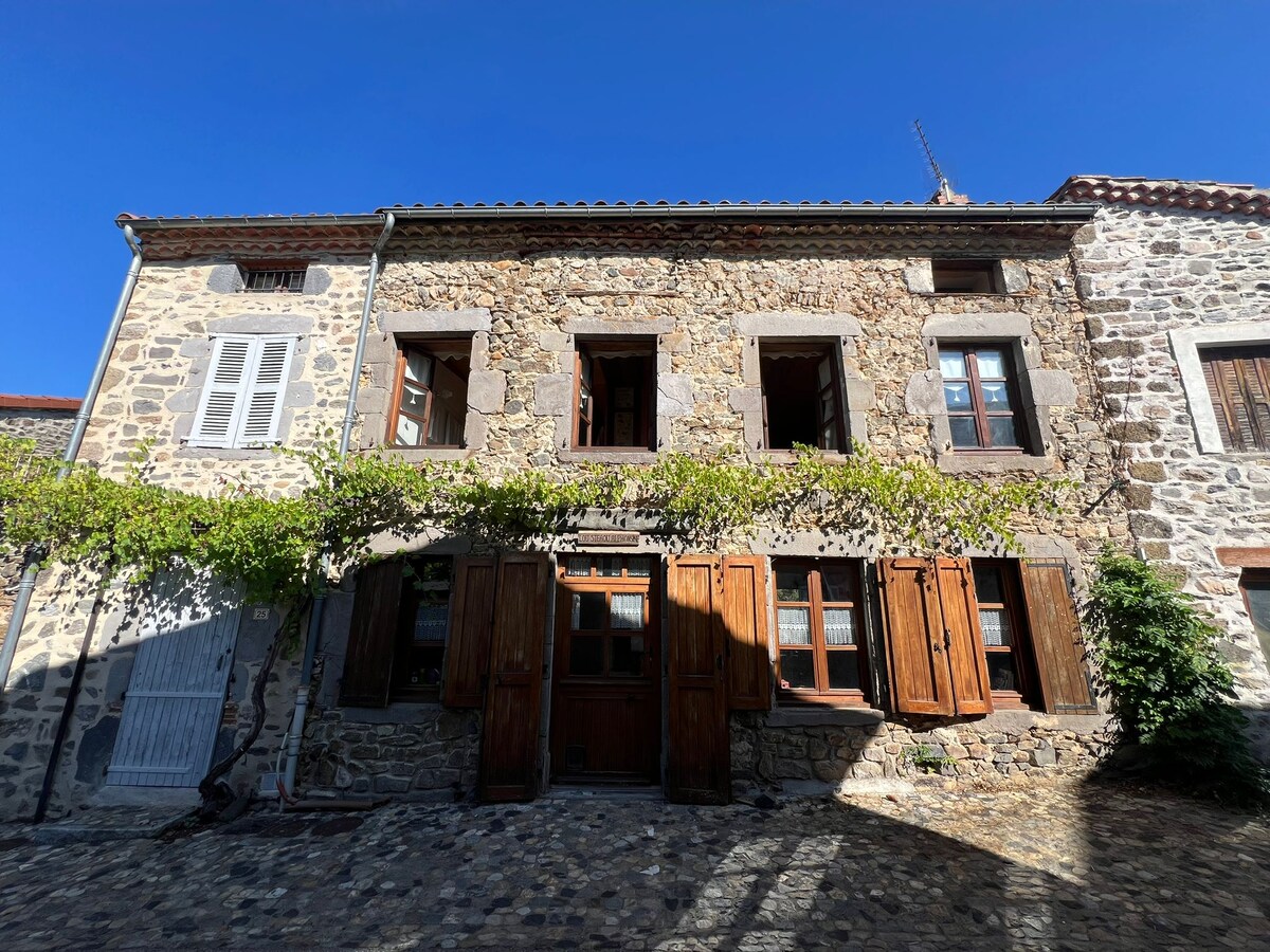 A charming stone house with wooden shutters stands prominently against a clear blue sky. The front façade features multiple windows flanked by climbing greenery, reflecting traditional architectural elements. Cobblestones lead up to the entrance, enhancing the rustic character of the home.