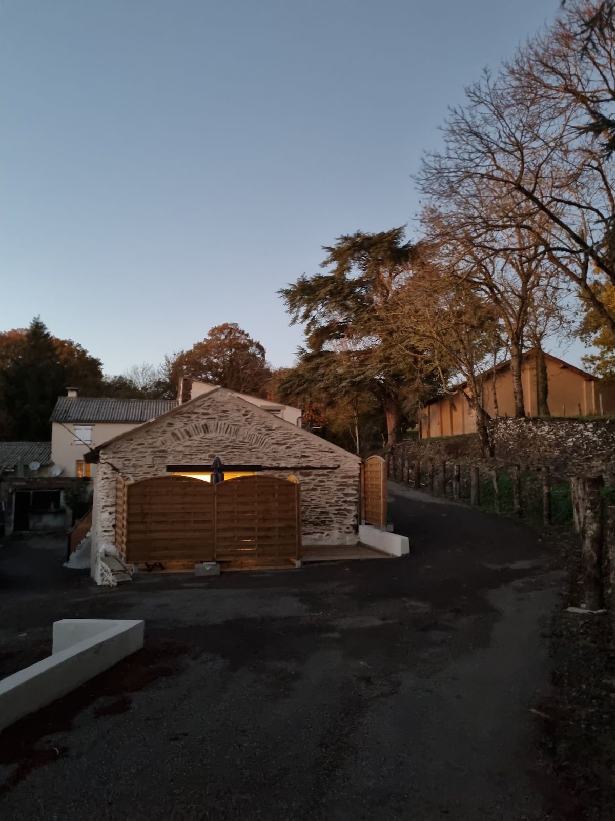 A serene exterior view of a stone cottage, illuminated softly at dusk. Surrounding trees display autumn foliage, while a gravel path leads to the building. The blend of natural and architectural elements creates a calm and inviting atmosphere in the countryside.