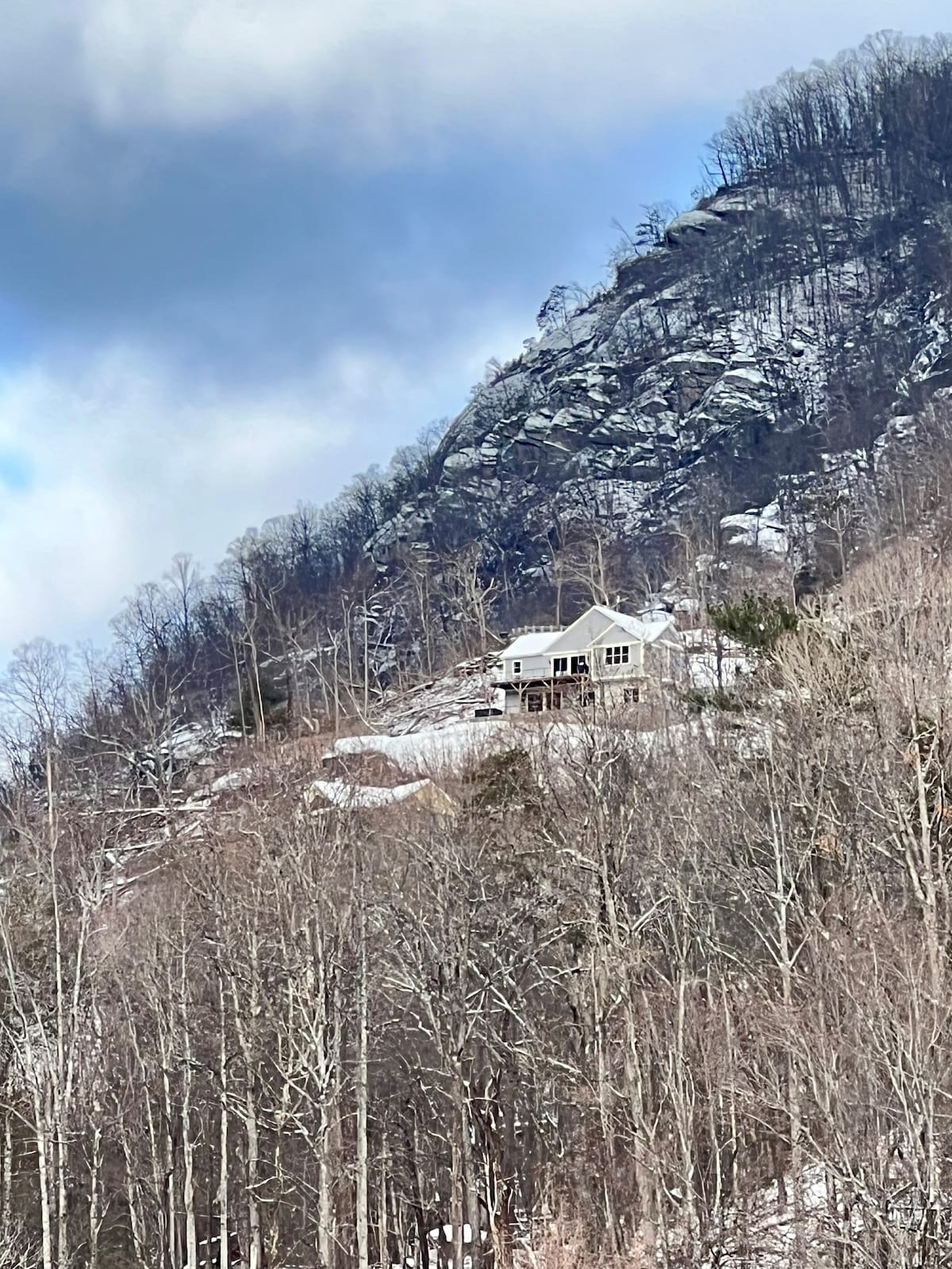 A mountain retreat is positioned on a hillside, surrounded by bare trees and rocky cliffs. The house features large windows that offer scenic views, all set against a backdrop of misty clouds and a rugged mountain landscape.