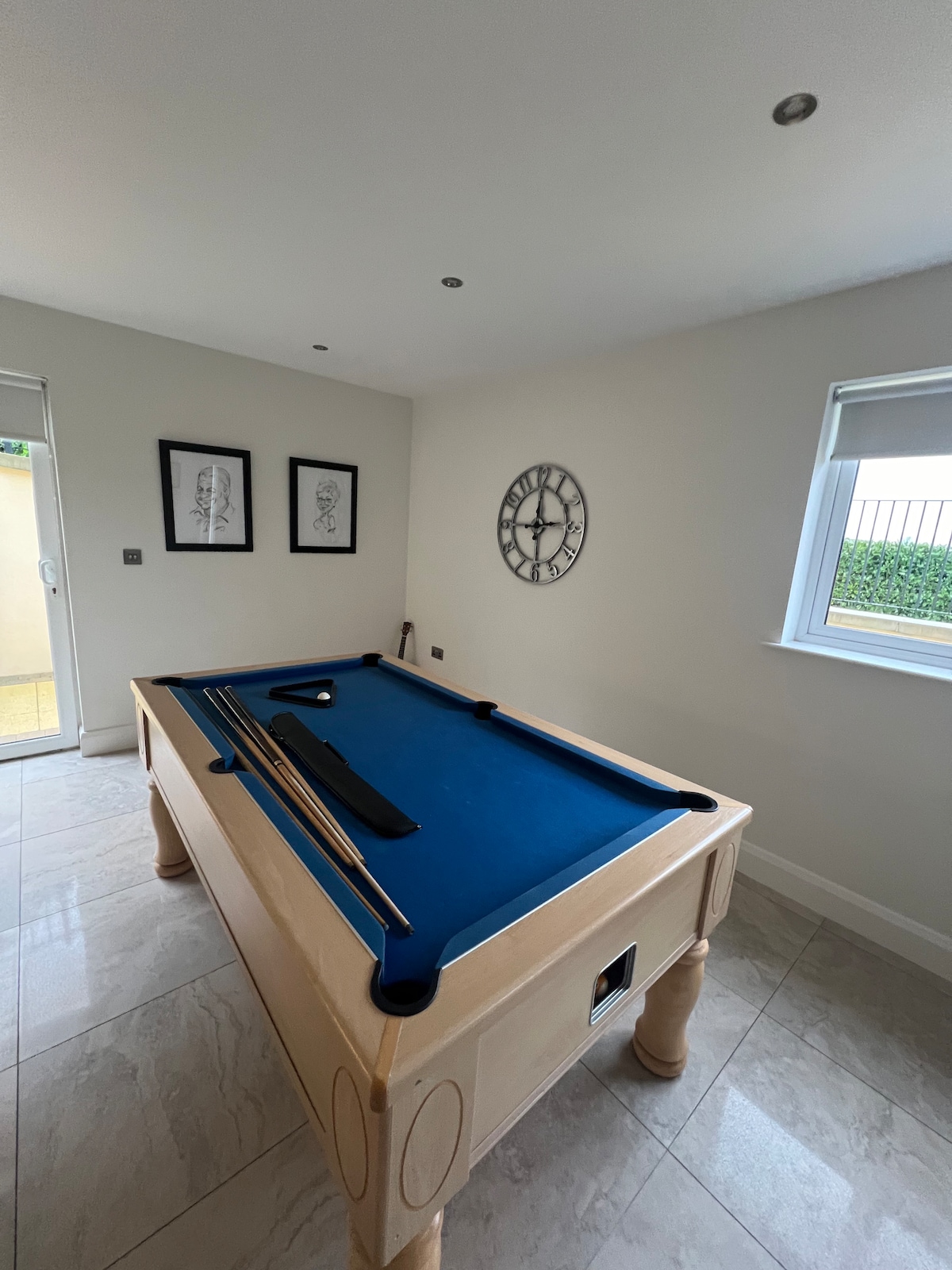 A bright and spacious game room is highlighted by a well-maintained pool table with a blue felt surface. Framed art pieces decorate the walls, while a large clock provides a focal point. Natural light enters through the window, enhancing the room's inviting atmosphere.