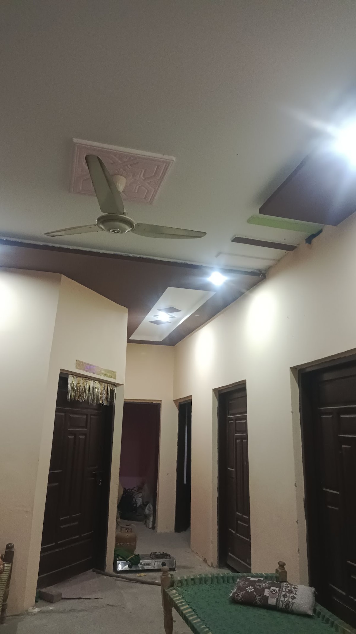 A well-lit corridor is lined with several closed doors, each featuring dark wooden panels. Soft lighting highlights the ceiling, which is adorned with a ceiling fan and decorative elements. The floor is unfinished, providing a glimpse of the ongoing improvements to the space.
