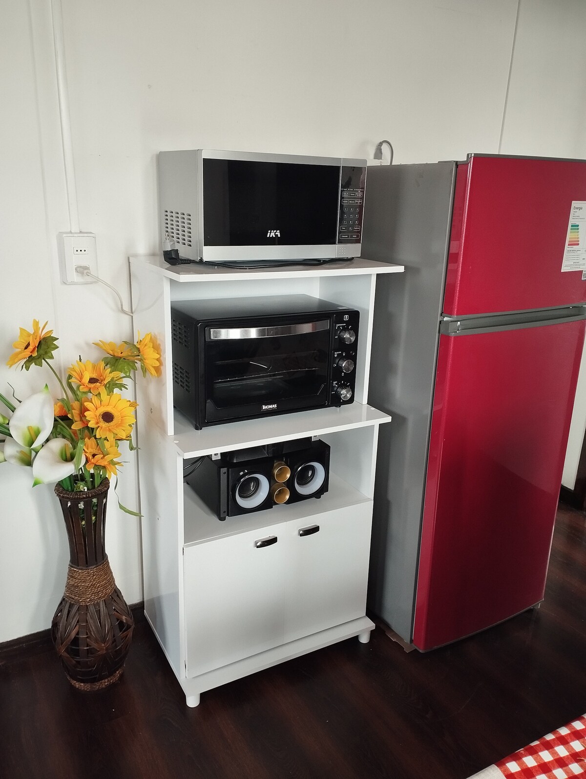 A well-organized kitchen area features a red refrigerator and a white shelf holding a microwave, an oven, and speakers. A decorative vase with sunflowers adds a touch of color to the space, contributing to a functional and inviting atmosphere.