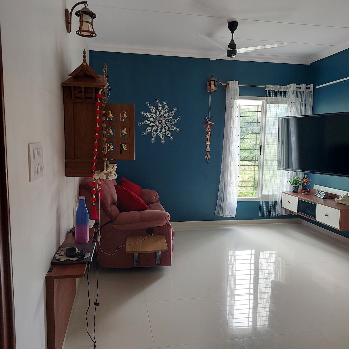 A living area is observed with a blue accent wall featuring decorative elements. A comfortable seating area with red cushions is positioned beside a wooden wall hanging. A flat-screen television is mounted opposite a window, allowing natural light to fill the space.