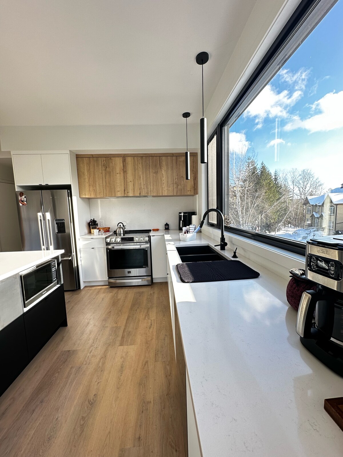 A modern kitchen features sleek cabinetry with wooden accents. Stainless steel appliances, including a refrigerator and stove, are visible. A large window allows natural light to fill the space, with views of the outdoors. A black sink is positioned next to a coffee maker and toasting equipment.