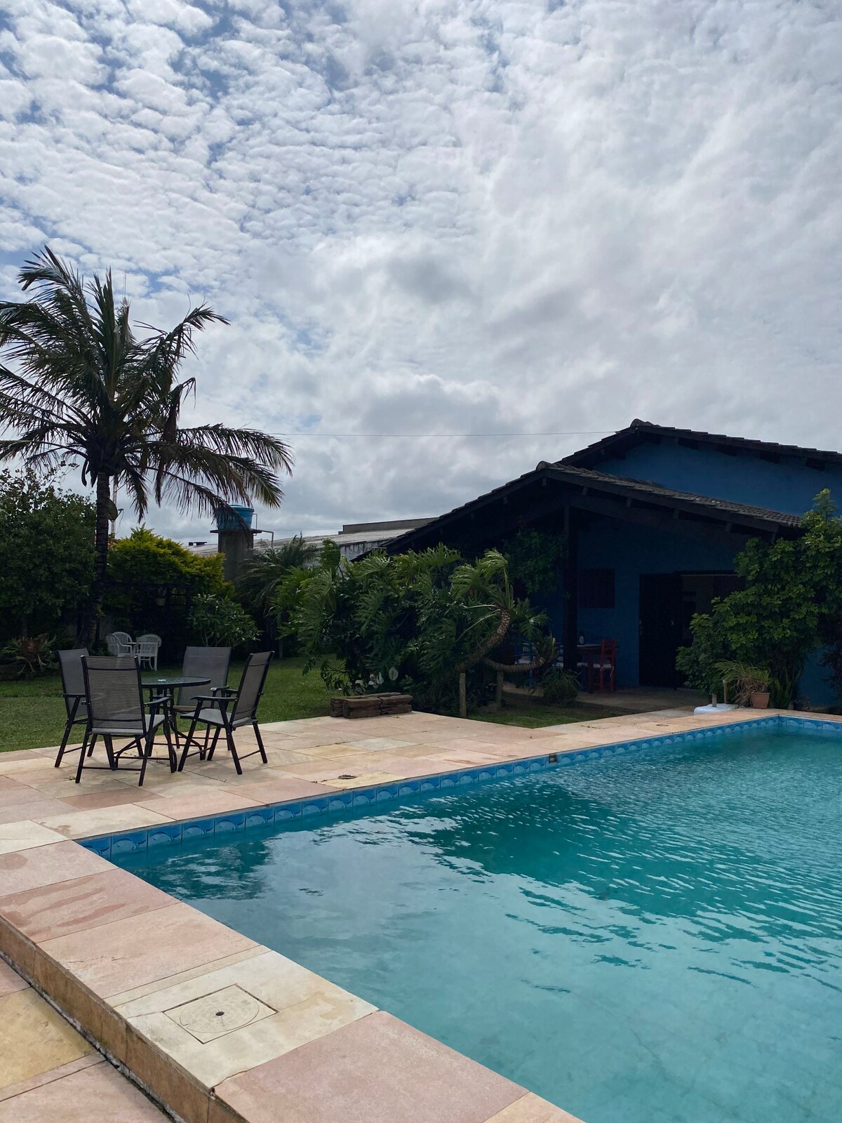 A swimming pool with a light blue hue is surrounded by a tiled patio. Nearby, a dining set features four black chairs and a table, complemented by lush greenery and palm trees in the background. A spacious house structure is visible, offering outdoor access.