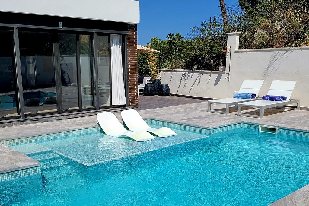 A bright outdoor area features a swimming pool with clear blue water, accompanied by two lounge chairs positioned beside it. Large glass doors allow for views of the interior, while the surrounding space includes minimalist landscaping and tiled decking.