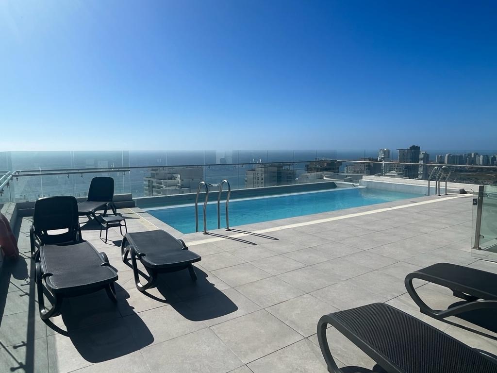 A rooftop pool area is showcased, featuring a clear blue water pool surrounded by sleek tiled flooring. Several lounge chairs are positioned nearby, providing an inviting space for relaxation while overlooking a panoramic view of the ocean and cityscape.