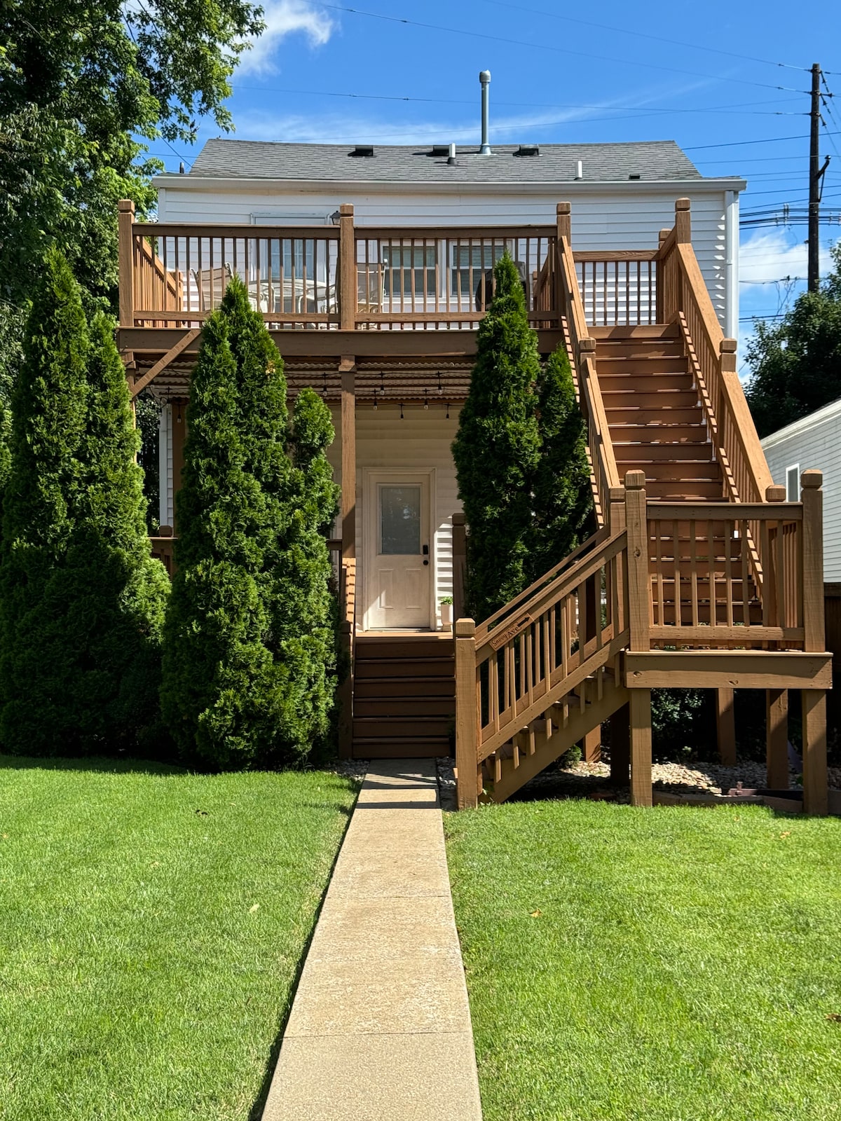 A charming exterior view features a wooden staircase leading to the second floor, flanked by tall green shrubs. A well-maintained lawn and paved walkway guide the entrance, which is brightened by natural light from above. The building's upper balcony offers a cooperative outdoor space.