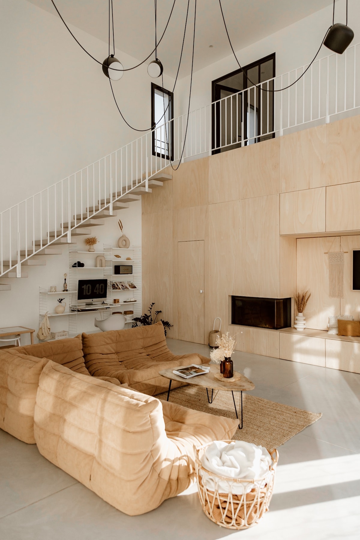 A spacious living area features a large, soft sofa in a neutral tone. A minimalist coffee table is placed in front, complemented by a decorative basket and a woven rug. The high ceiling is adorned with modern light fixtures, enhancing the area’s open feel.