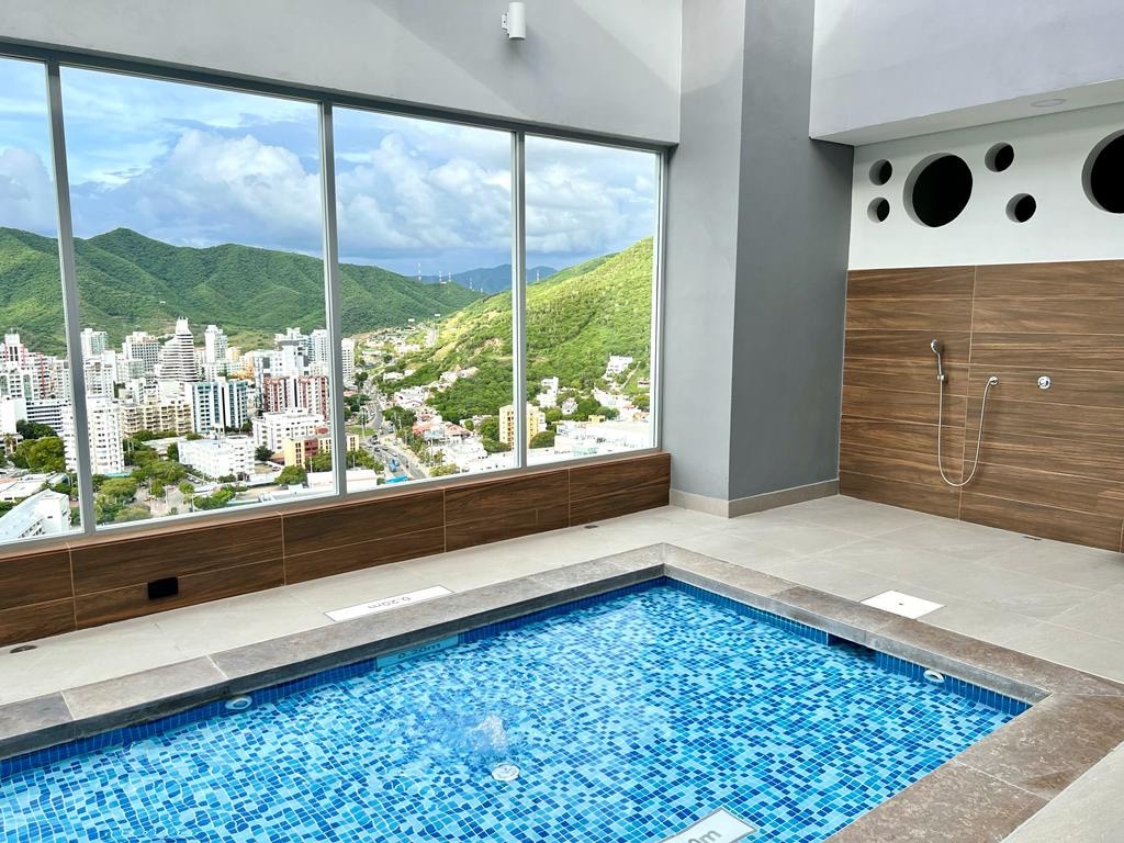 A bright area features a small, inviting pool with a mosaic tile design. Large windows showcase a panoramic view of the surrounding mountains and cityscape. Natural light fills the space, complemented by neutral-toned walls and wooden accents.