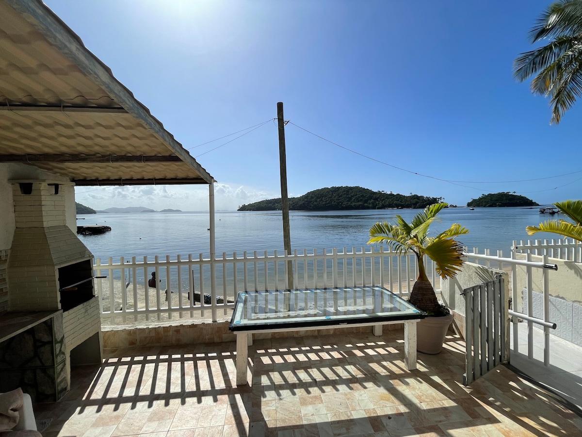 A scenic outdoor patio is depicted, featuring a glass table surrounded by a mix of tropical plants. The view includes a tranquil body of water, with distant green islands visible against a clear blue sky, complemented by a hint of sunlight reflecting off the water.