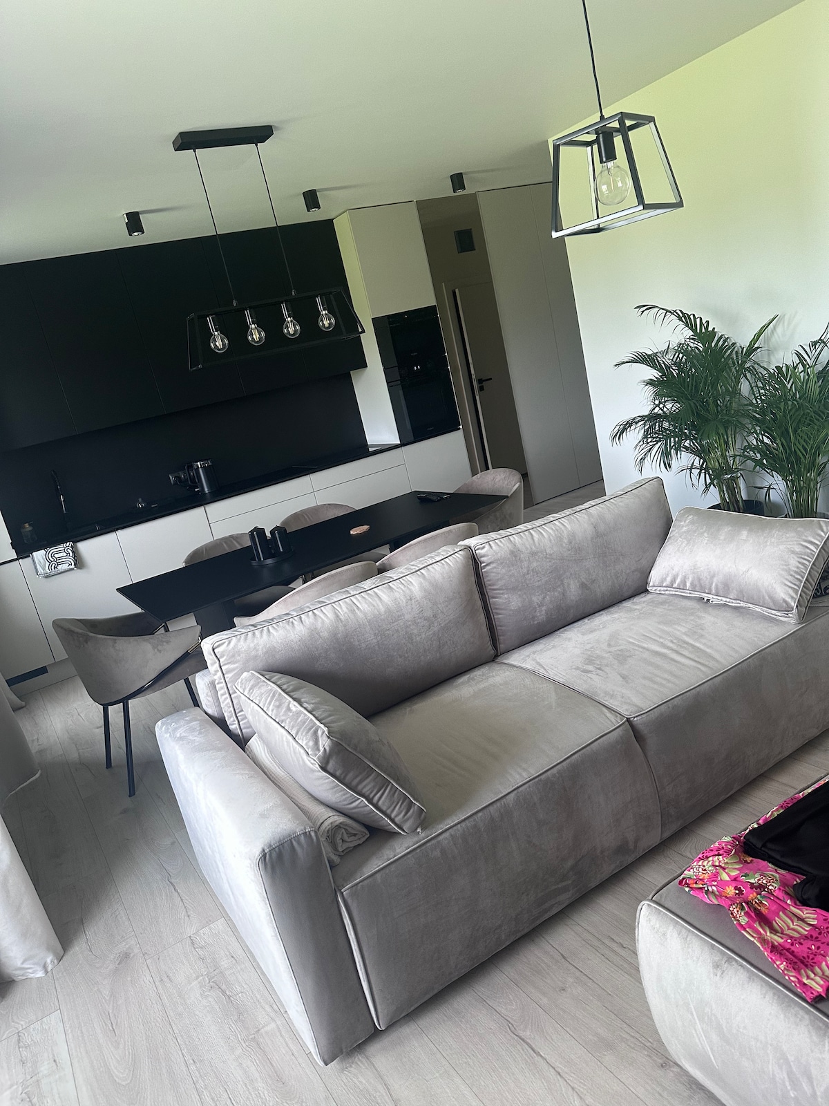 A modern living area features a sleek, light-colored sofa facing a stylish dining table set for four. A geometric pendant lamp hangs overhead, complementing the minimalist decor. In the background, a tall indoor plant adds a touch of greenery to the space.