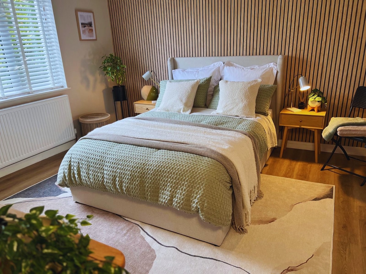 A spacious bedroom features a comfortable bed adorned with soft pillows and a textured blanket. Nightstands with lamps sit on either side, complemented by a cozy chair and decorative plants. Natural light fills the room through the window covered by wooden blinds.