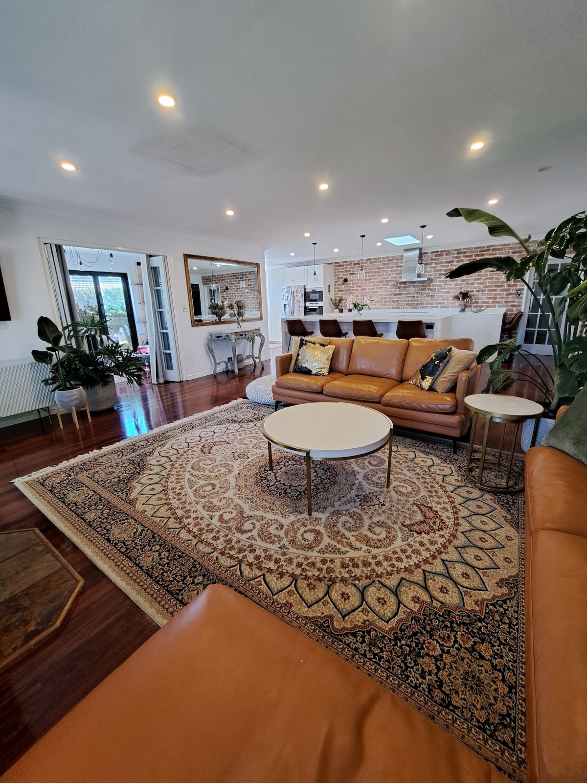 The open-plan living area features a warm leather sofa, accent pillows, and a decorative area rug. A round coffee table is centered amidst the seating. Light streams in through large windows, highlighting the stylish brick wall and connecting to the kitchen area in the background.