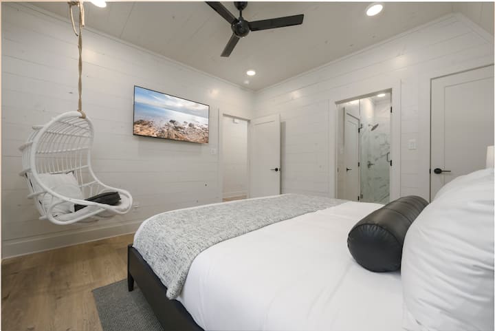 Luxury king bedroom with white bedding, 55” HD smart TV, hanging chair and attached bathroom.