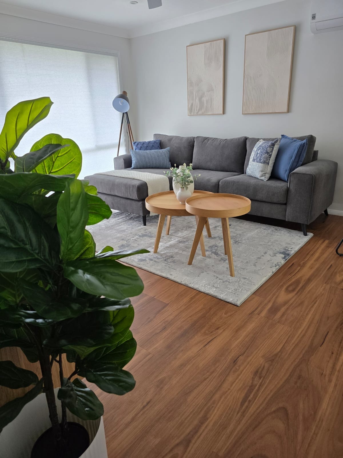 A cozy living area features a grey sectional sofa adorned with decorative pillows. Light-colored wooden side tables are positioned in front of the sofa, complemented by a textured area rug. A leafy plant is visible in the foreground, adding greenery to the space.