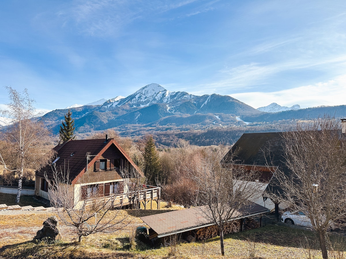 The view reveals a serene mountainous landscape, featuring snow-capped peaks under a clear blue sky. Two wooden structures are positioned in the foreground, surrounded by a grassy area dotted with trees. The peaceful surroundings highlight the natural beauty of the location.