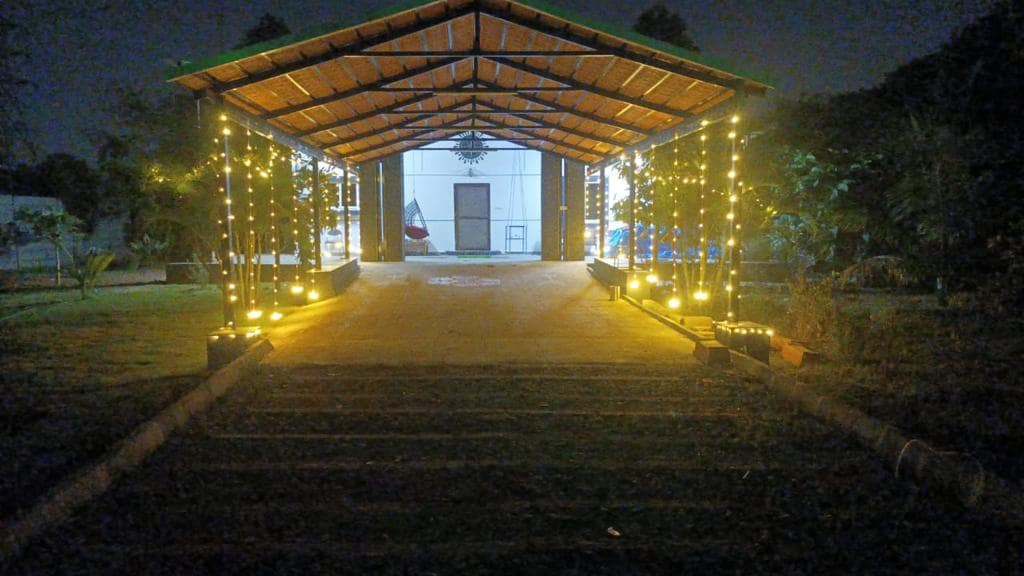 An outdoor entrance is adorned with warm lights, highlighting the pathway leading to a covered structure. Lush greenery surrounds the area, creating a serene atmosphere. The roof features a wooden design, enhancing the rustic charm of the setting, while soft lighting casts a welcoming glow.
