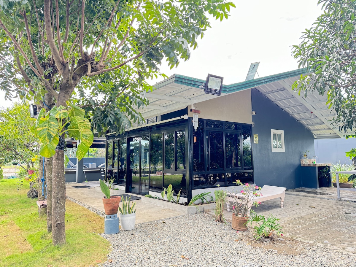 A black-framed house features large glass doors that provide views of the surrounding greenery. Potted plants are arranged along the pathways. An outdoor seating area is visible, under a covered porch that enhances the outdoor living experience.