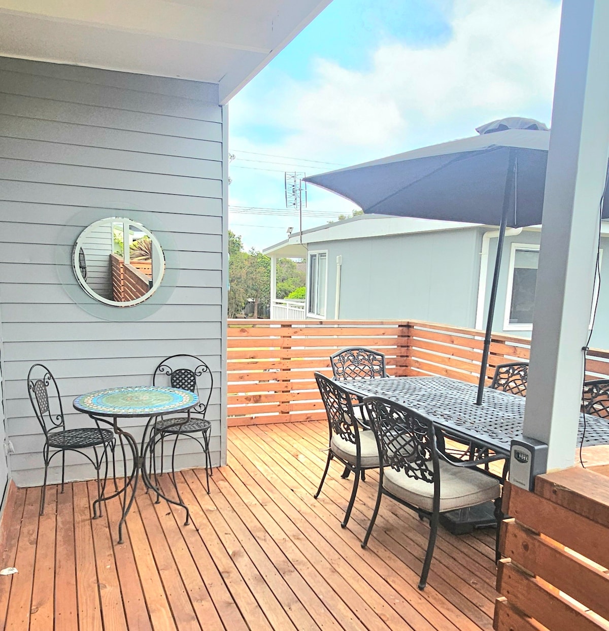 A sunlit deck area features a small round table with two chairs, alongside a larger dining table accommodating six. A large umbrella offers shade, and a mirror reflects the inviting outdoor space. Wooden fencing provides some privacy, creating a cozy spot for relaxation.