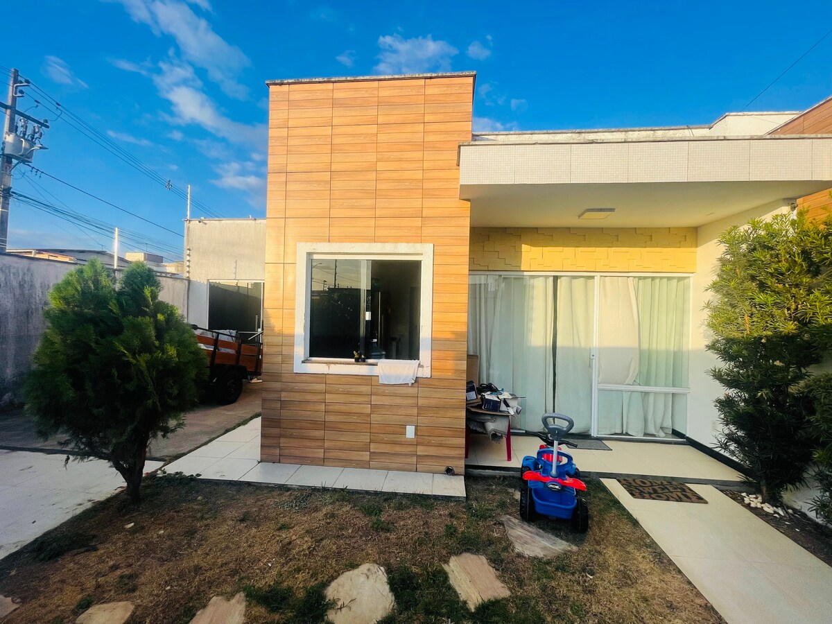 A modern corner house is presented with a warm wood-textured exterior. Large glass windows provide ample natural light, while sheer curtains gently filter it. A small, manicured lawn is situated in front, featuring a playful children's ride-on toy and bushes lining the pathway.