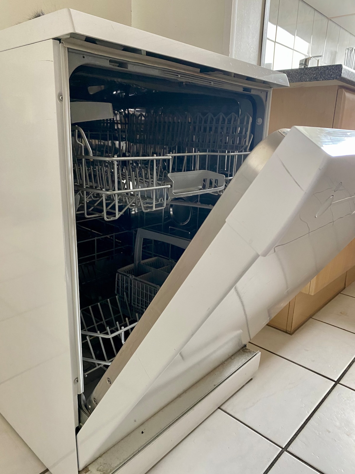 A dishwasher is shown partially opened, displaying its spacious interior equipped with multiple racks for dishes. The exterior is clean and white, complementing the surrounding kitchen cabinetry. The floor features light-colored tiles, enhancing the overall modern aesthetic.