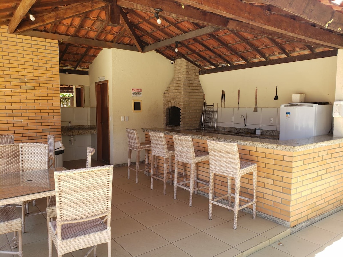 An outdoor dining area features a large counter with bar stools, complemented by a stone grill and a spacious kitchen space. A tiled floor extends throughout, while natural light fills the area through an open design beneath a wooden roof.