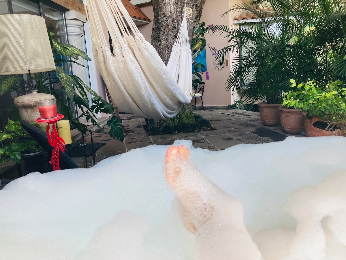 Bubbles cover the surface of a jacuzzi, partially obscuring the view of feet submerged in the water. Surrounding greenery includes potted plants and a hammock stretched between two trees, creating a peaceful outdoor retreat.