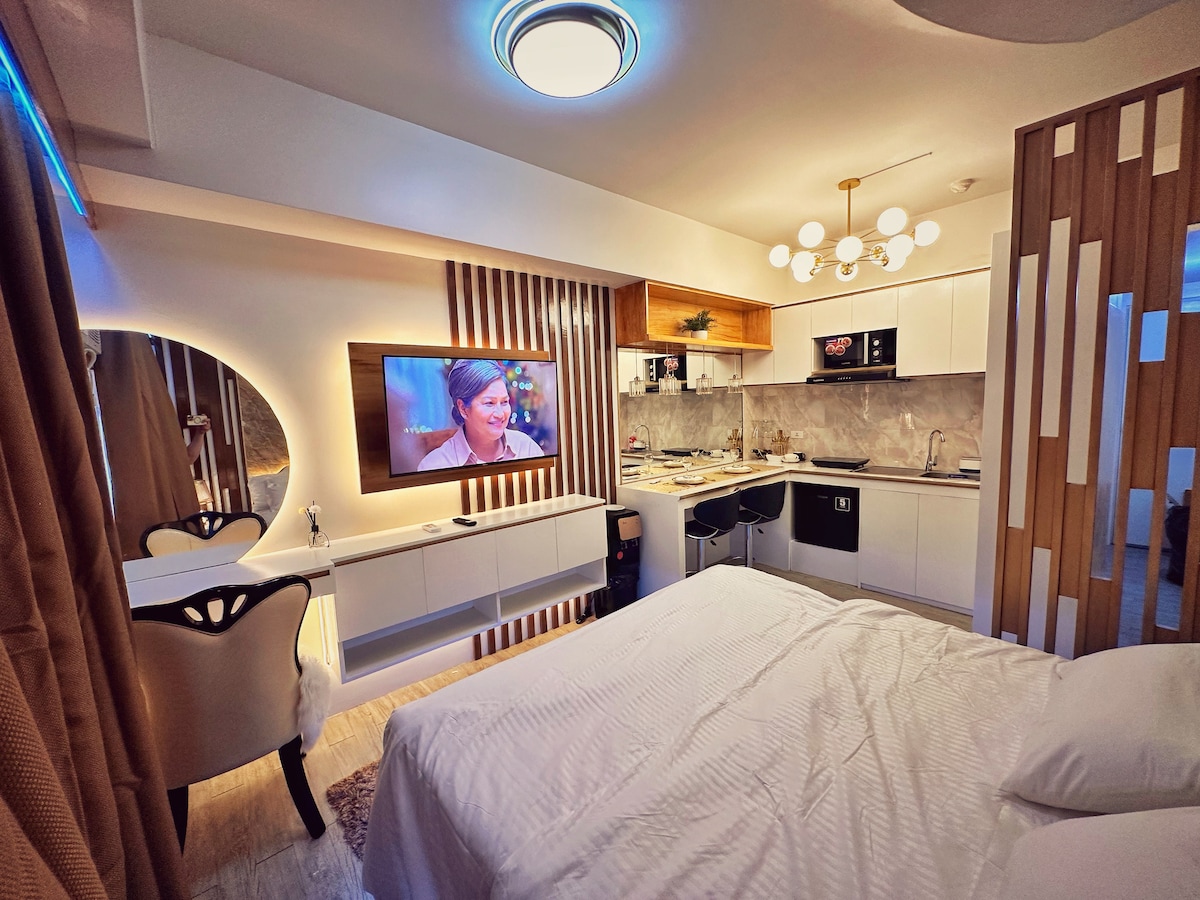 A modern studio apartment features a spacious bed with crisp white linens, complemented by sleek wooden accents. A wall-mounted television is positioned above a minimalist kitchen area, which includes essential appliances. Soft lighting from a stylish ceiling fixture and warm tones enhance the contemporary design.