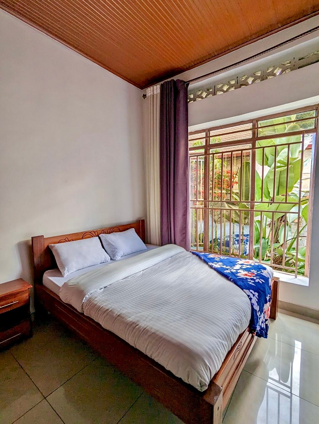 A cozy sleeping area features a wooden bed with a neatly arranged white bedspread and two pillows. Natural light filters through the window, highlighting the greenery outside. Light-colored walls contrast with the warm wooden ceiling, creating a calming atmosphere.