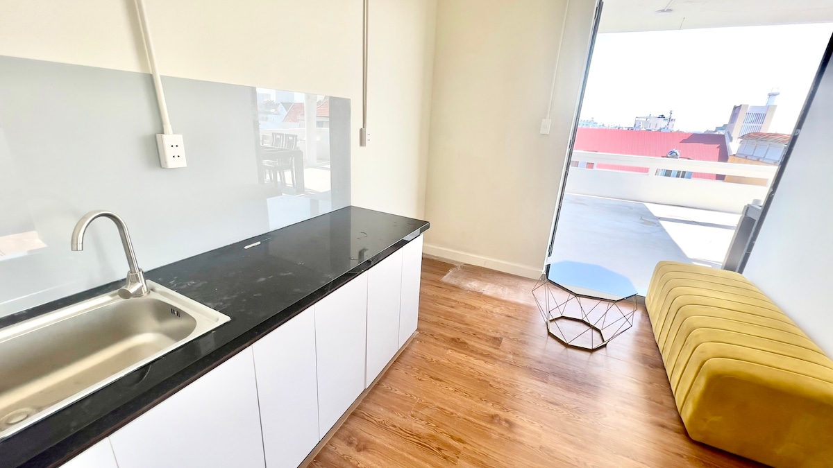 A tidy kitchen area features a stainless steel sink set into a black countertop. White cabinetry complements the space, while a golden sofa and a geometric metal side table add warmth. A bright balcony is visible through sliding glass doors.