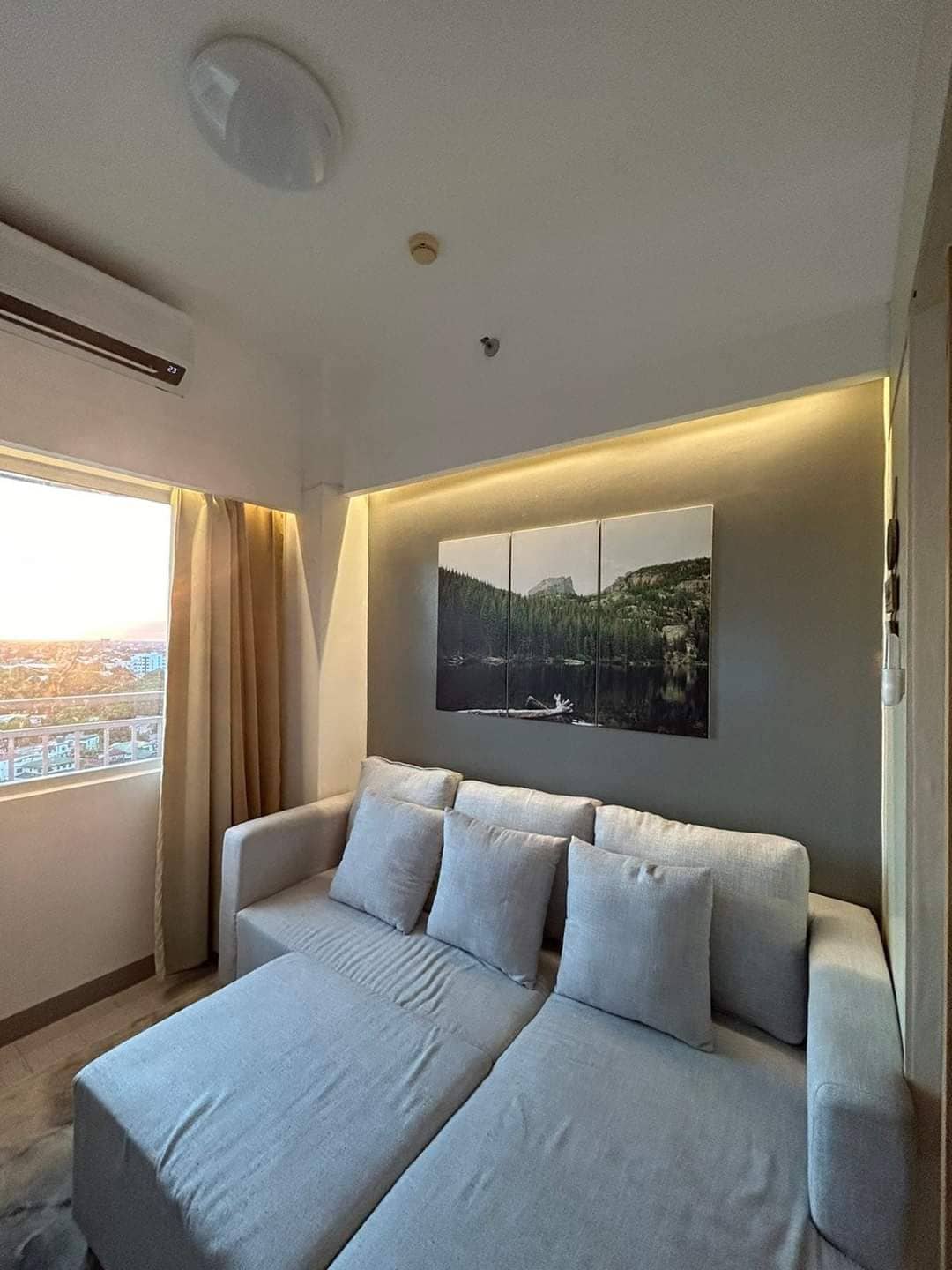 A light-filled living area features a comfortable sofa with several cushions. A large landscape painting depicting a serene natural scene adorns the wall above. Floor-to-ceiling curtains frame a window offering views of the outside, enhancing the peaceful ambiance.