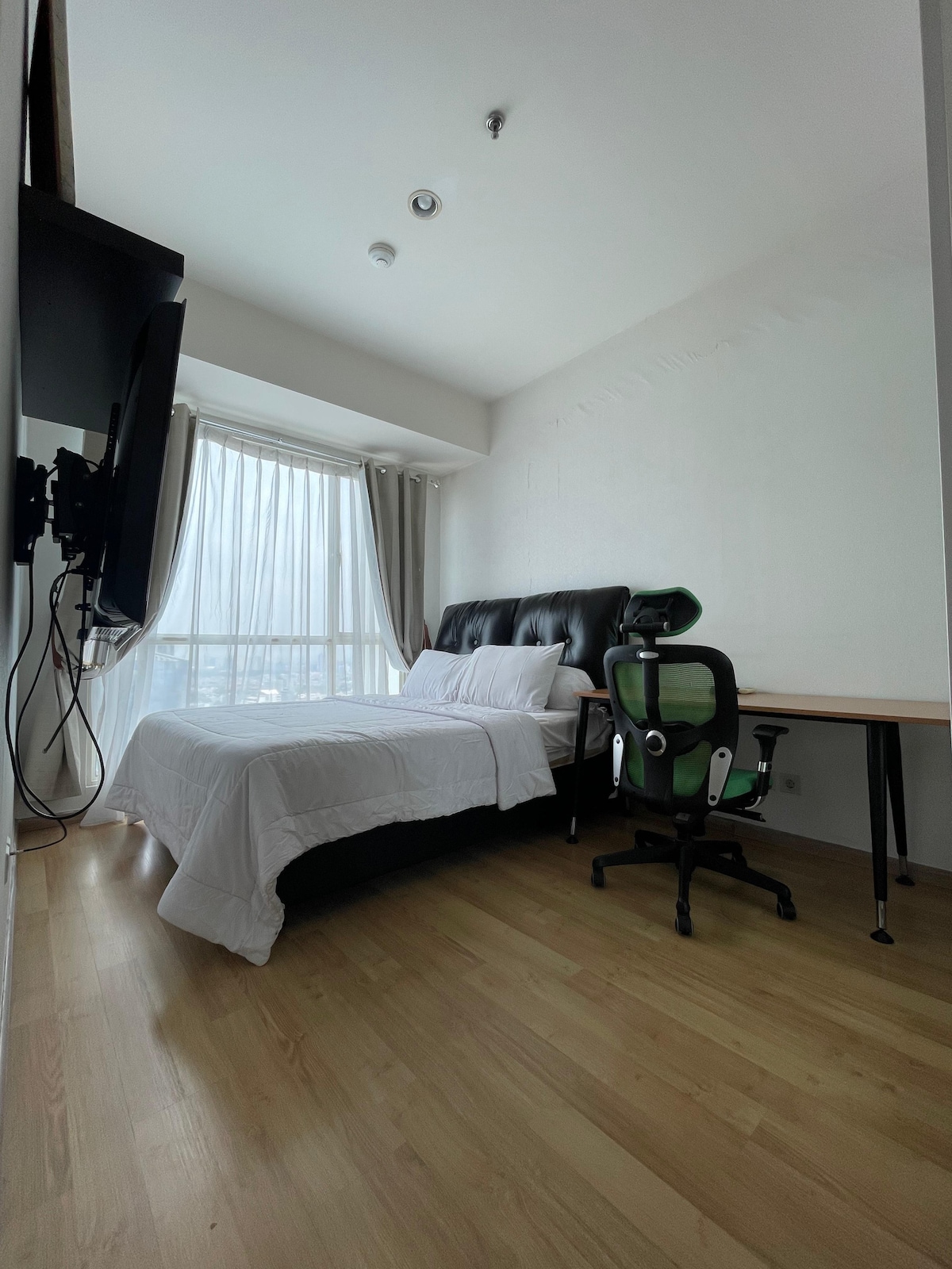 A well-lit bedroom features a spacious bed with a dark headboard and crisp white linens. A large window allows natural light to flow in, complemented by light curtains. A desk and ergonomic chair are included, providing a functional workspace.